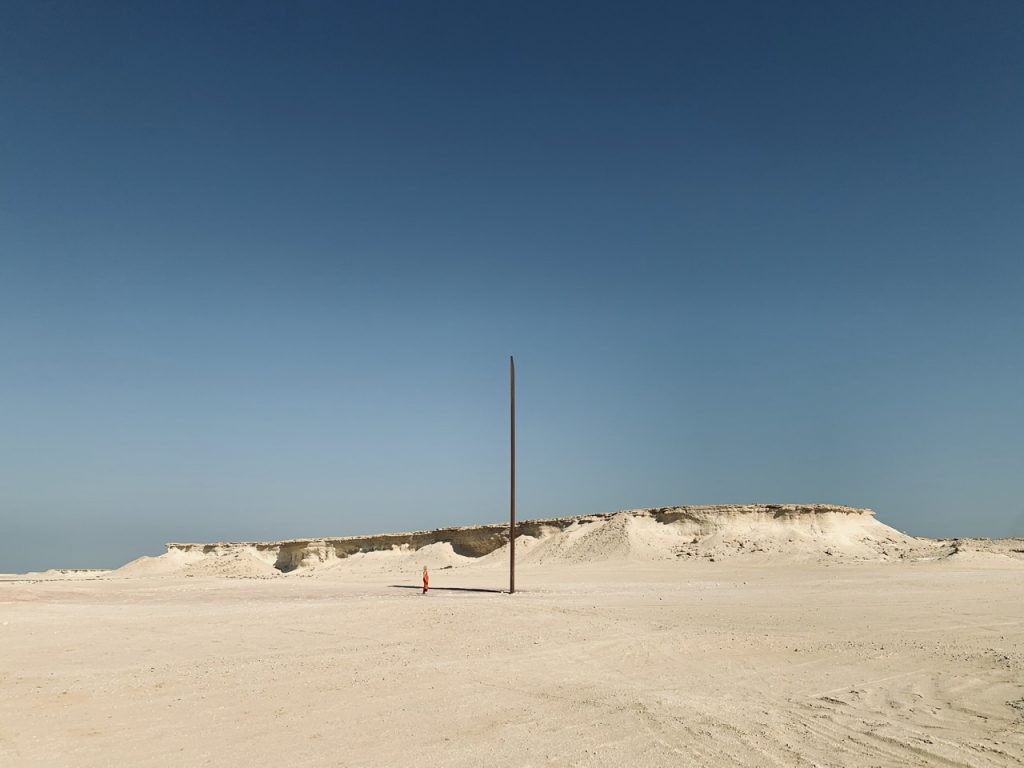 Large steel Richard Serra’s sculpture, Tijana in a burnt orange Kalita dress, Doha desert skyline. Qatar.