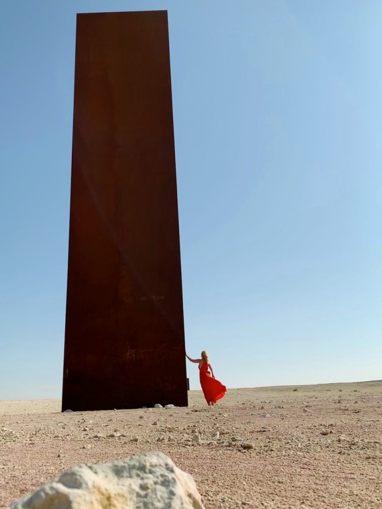 Tijana in an orange Kalita dress standing by oxidized steel sculpture by Richard Serra in Qatar.

