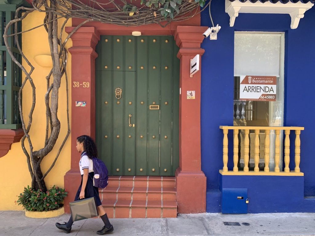 A young girl walks to school in front of bright yellow, brick, and blue colored walls in Cartagena