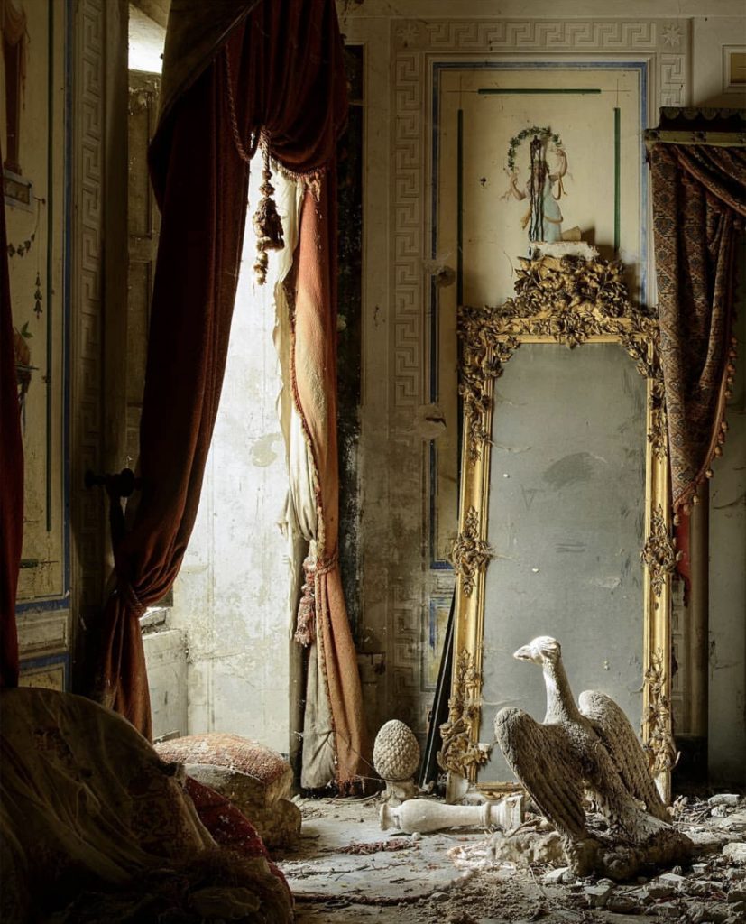 L'Horizon Chimérique by Nicola Bertellotti. Light pours through decaying curtains onto a dusty but intricate gold mirror and bust of an eagle. 